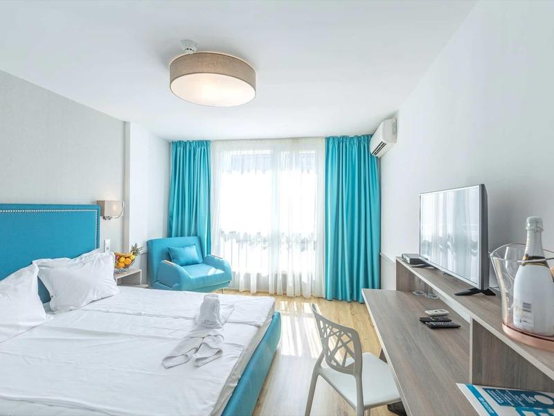 Lichte hotelkamer met tweepersoonsbed, turquoise gordijnen, fauteuil en bureau met tv.