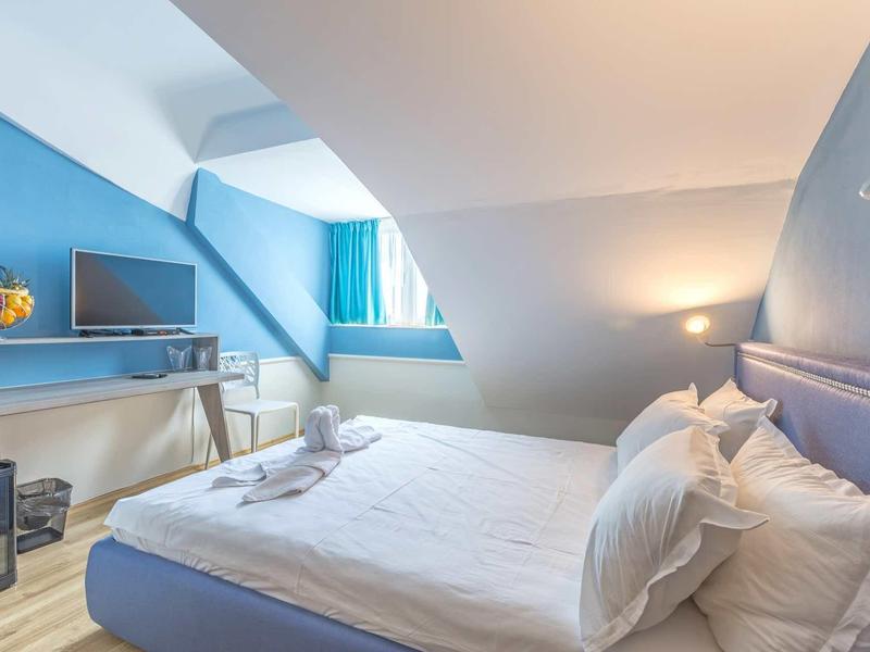 Modern hotelkamer met blauw bed, bureau en tv onder schuine muur.