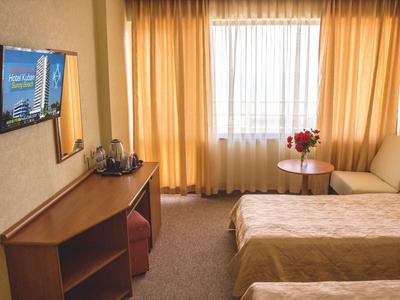 Bright hotel room with two single beds, sofa, table with flowers, and wall-mounted TV.