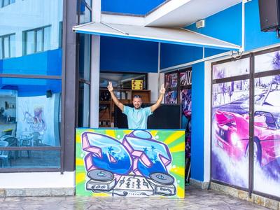 A man waves behind a colorful reception desk in front of a blue building.