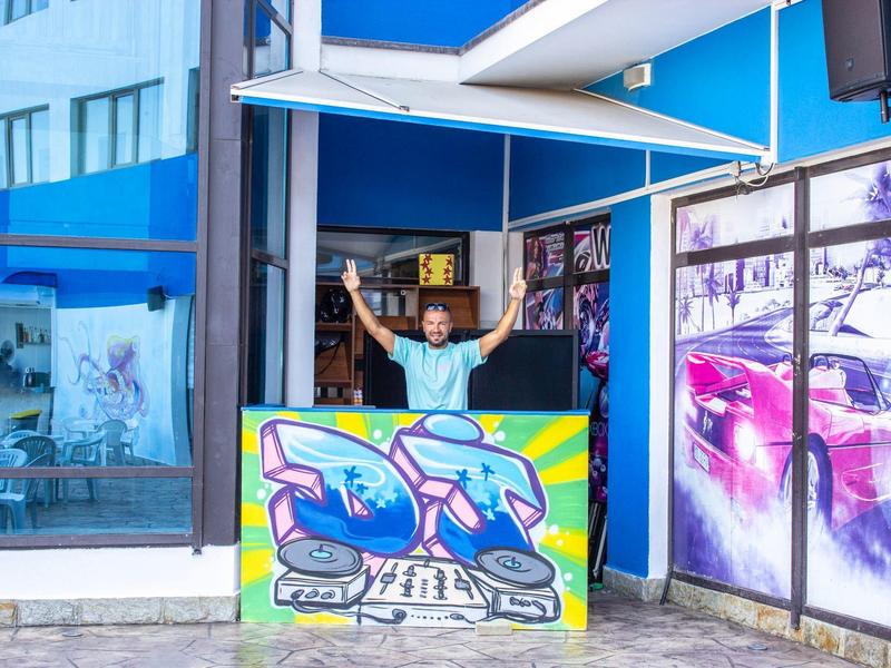 A man waves behind a colorful reception desk in front of a blue building.