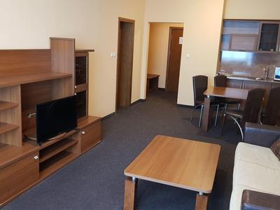 Moderne hotelsuite met houten meubels, tv, bank, eettafel en kitchenette in neutrale kleuren.