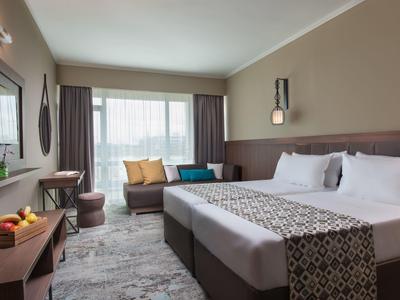 Moderne hotelkamer met groot tweepersoonsbed, bank, bureau, natuurlijk licht en warme kleuren.