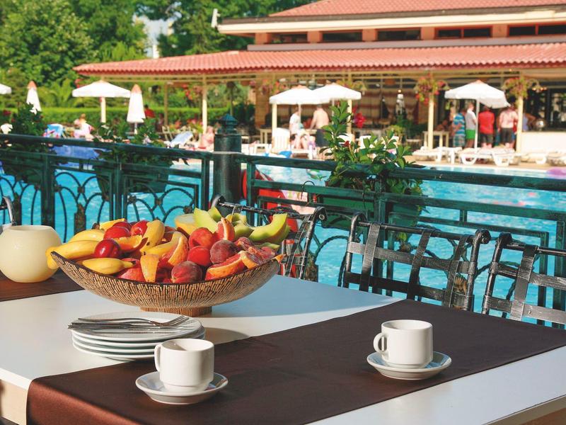 Tafel met koffiekopjes en fruitschaal naast zwembad en parasols in hotel tuin.