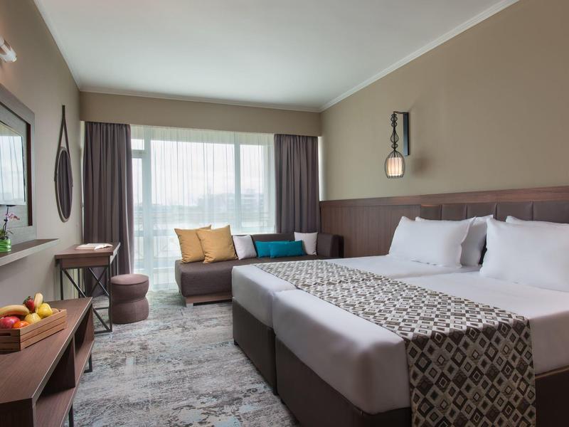 Moderne hotelkamer met groot tweepersoonsbed, bank, bureau, natuurlijk licht en warme kleuren.