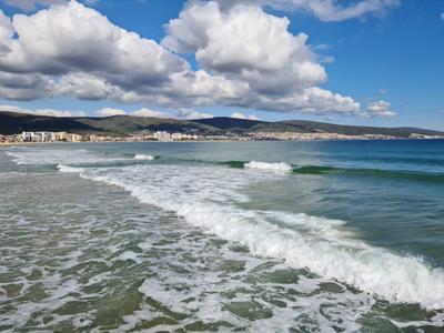 Golven slaan tegen een zandstrand met een bewolkte hemel en heuvels op de achtergrond.
