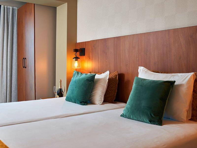 Moderne hotelkamer met twee eenpersoonsbedden, groene kussens en warme houten panelen achter het bed.