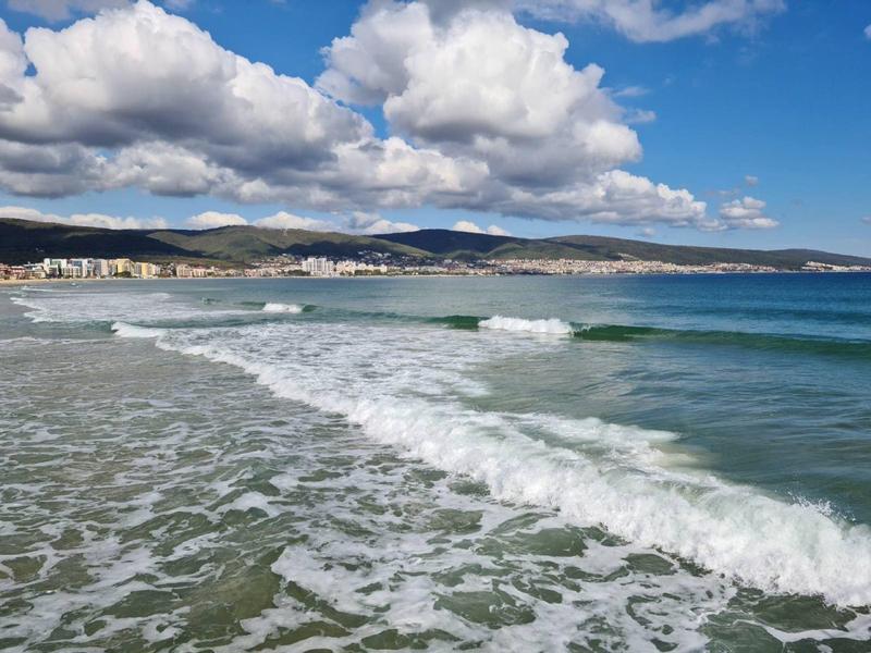 Golven slaan tegen een zandstrand met een bewolkte hemel en heuvels op de achtergrond.