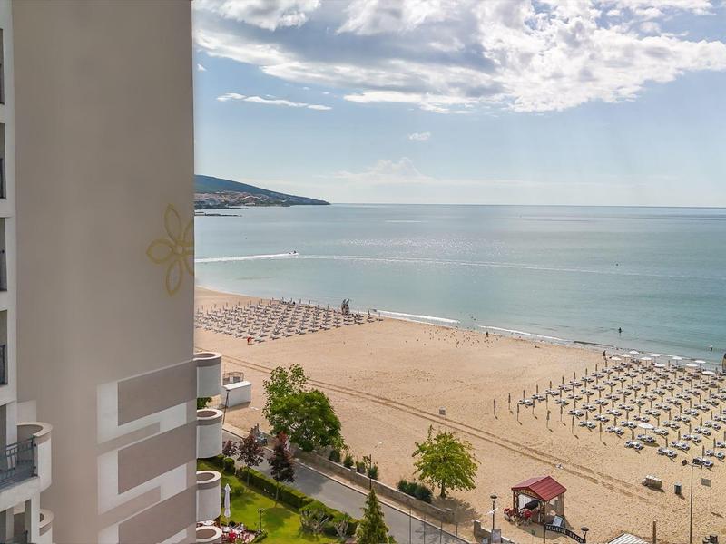 Uitzicht op een zandstrand met ligbedden en een hotelgebouw onder een zonnige hemel.