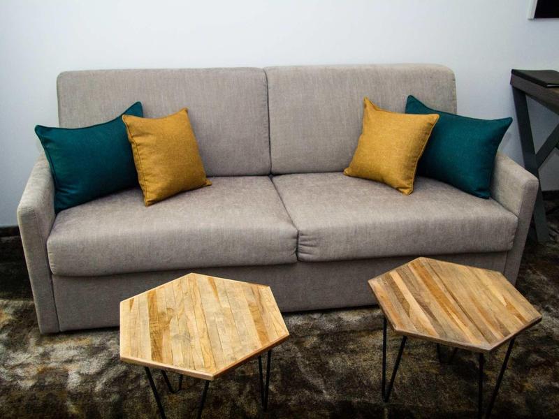 Beige bank met groene en gele kussens voor twee zeshoekige houten tafels in een kamer.