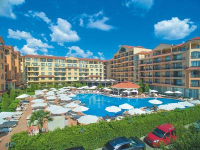 Groot hotel met buitenzwembad, ligstoelen en parasols onder een blauwe lucht.