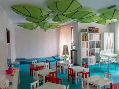 Kleurrijke speelruimte met kleine tafels en stoelen, groene bladeren aan het plafond en een boekenplank.