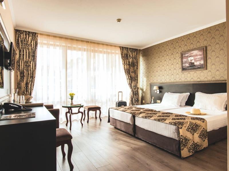 Lichte hotelkamer met twee bedden, bureau, stoel en grote ramen met gordijnen.