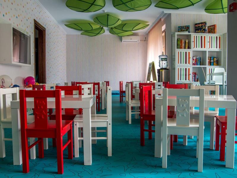 Kinderkamer met kleurrijke tafels en stoelen, groene bladddecoraties aan het plafond, boekenplank op de achtergrond