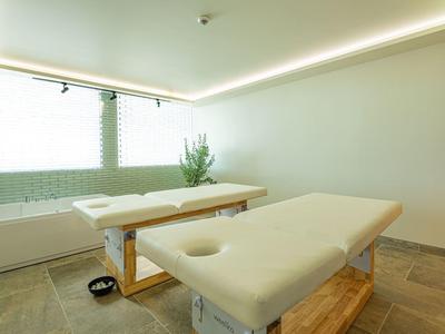 Lichte behandelruimte met twee massagetafels en groene tegels bij het raam.