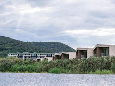 Moderne gebouwen aan een meer met een beboste heuvel op de achtergrond.