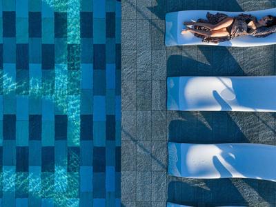 Vier weiße Liegestühle neben einem blauen Schwimmbecken auf einer Steinfliesenfläche.