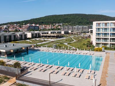 Moderner Hotelkomplex mit großem Außenpool, Liegestühlen und grünen Rasenflächen bei sonnigem Wetter.