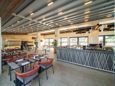 Lichte, moderne restaurantruimte met stoelen, tafels, grote ramen en een lange bar.