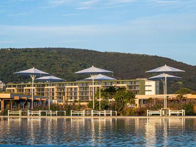 Modernes Hotel mit Pool und Sonnenschirmen vor bewaldetem Hügel unter blauem Himmel