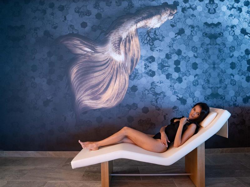 Vrouw ontspant op ligstoel voor wand met vispatroon in hotelkamer.