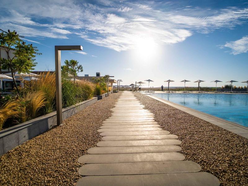 Een zonnig pad naast een zwembad leidt naar een rij parasols aan het strand.