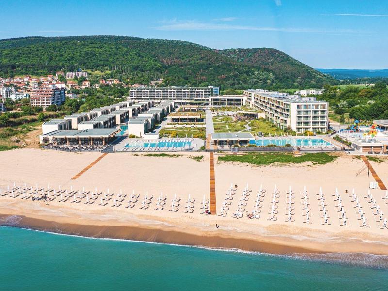 Breed zandstrand met grote hotels, zwembad en uitzicht op heuvels en zee.