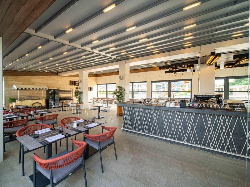 Lichte, moderne restaurantruimte met stoelen, tafels, grote ramen en een lange bar.