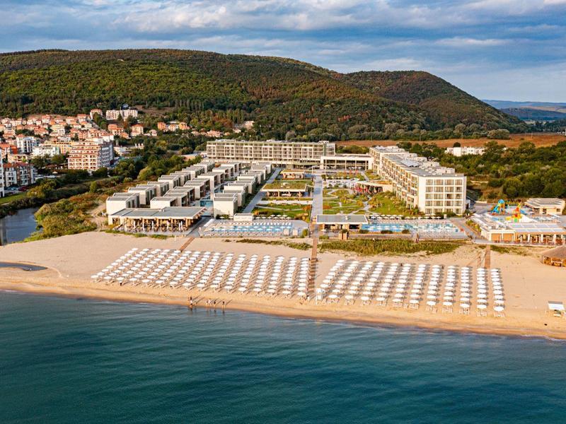 Strand met ligstoelen en een hotel aan de voet van een beboste heuvel op een heldere dag.