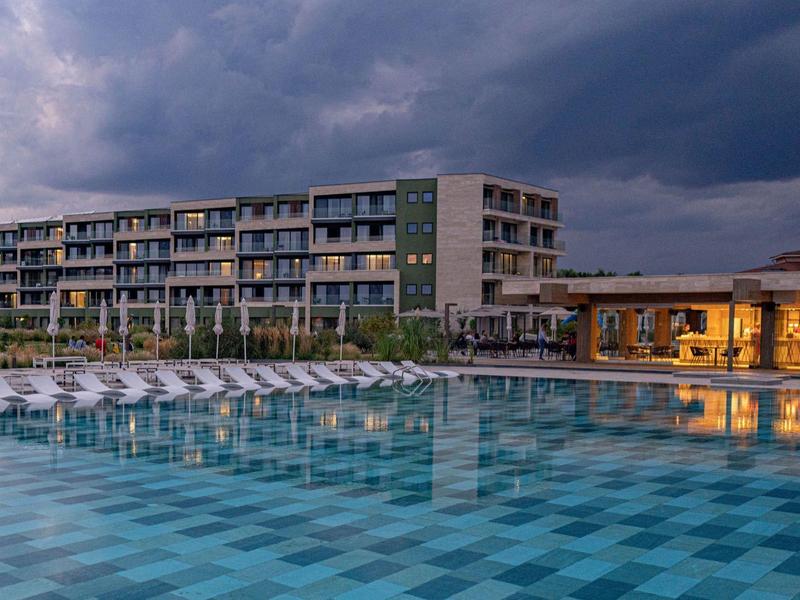 Modern hotel met groot zwembad en ligstoelen onder bewolkte lucht.