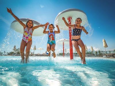 Drei Kinder springen fröhlich in einen Pool bei sonnigem Himmel und Wasserspielen.