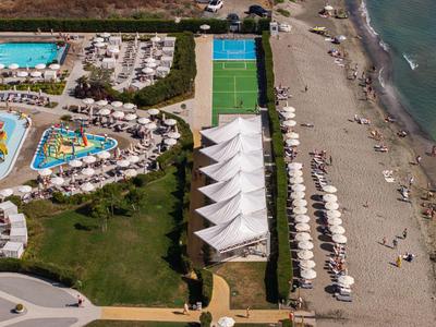 Luftbild eines Resorts mit Pool, Wasserrutschen, Tennisplatz und Sandstrand mit Sonnenschirmen.