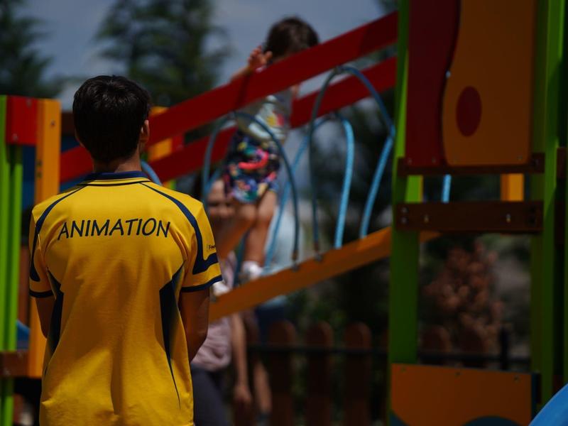Ein Betreuer in gelbem Animation-Shirt beobachtet Kinder auf einem bunten Spielplatz.