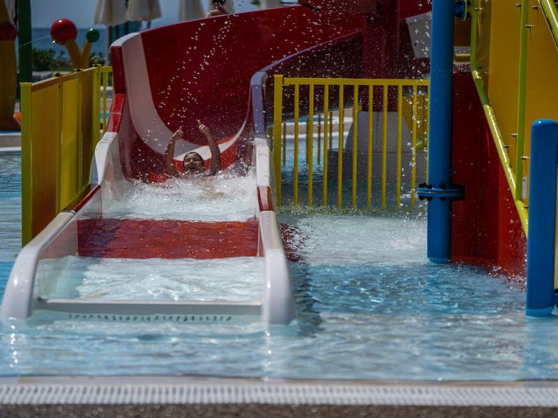 Rote Wasserrutsche mündet in ein flaches Schwimmbecken mit buntem Geländer.