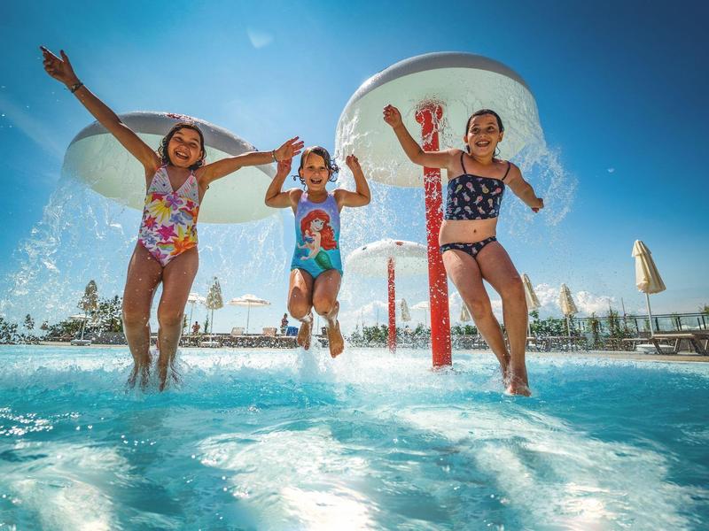 Drei Kinder springen fröhlich in einen Pool bei sonnigem Himmel und Wasserspielen.