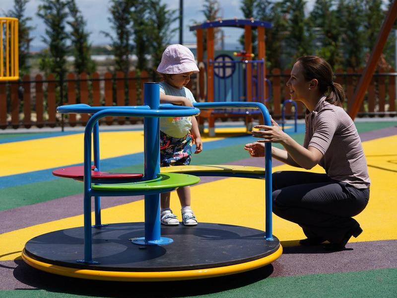 Ein Kind auf einem Karussell im Park, betreut von einer Frau auf farbigem Boden.