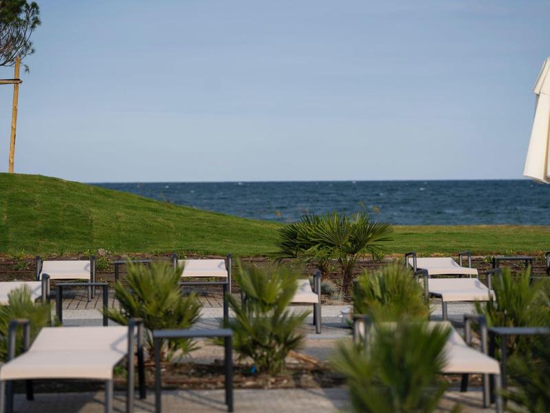Leere Sonnenliegen mit weißen Auflagen und geschlossener Sonnenschirm am Meerblick unter blauem Himmel