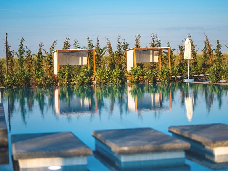 Moderner Pool mit Liegen und Pavillons vor grünem Hain bei blauem Himmel.