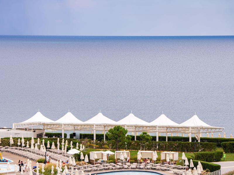 Blick auf einen Poolbereich mit Sonnenschirmen vor ruhigem Meer und blauem Himmel.