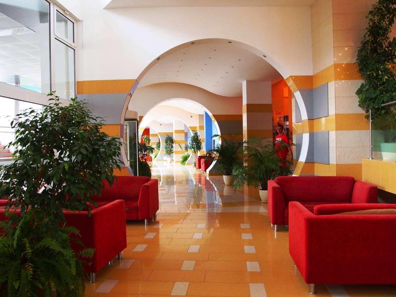 Bright hotel lobby with red armchairs, plants, and arches in the background.