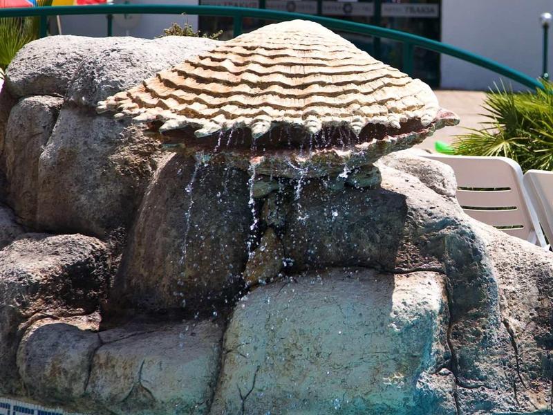 Large rock with artificial waterfall next to blue pool in a hotel area.