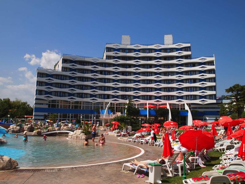 Modern hotel with wave-shaped glass facade and outdoor pool with sun loungers and red umbrellas.