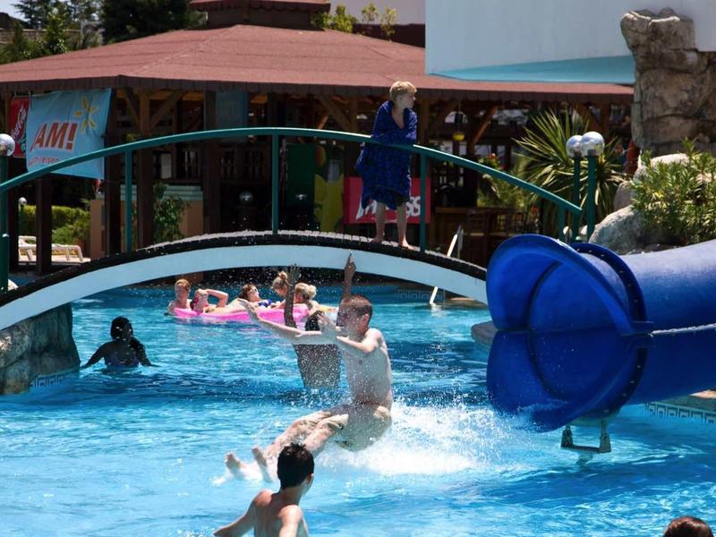 People swim and slide in a pool with a blue water slide and a bridge.