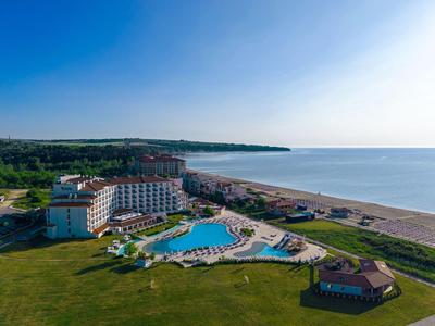 Grande hotel con piscine e spiaggia sulla costa, circondato da verde e cielo azzurro.