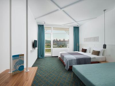 Camera d'hotel moderna con tre letti, moquette verde e balcone con vista.