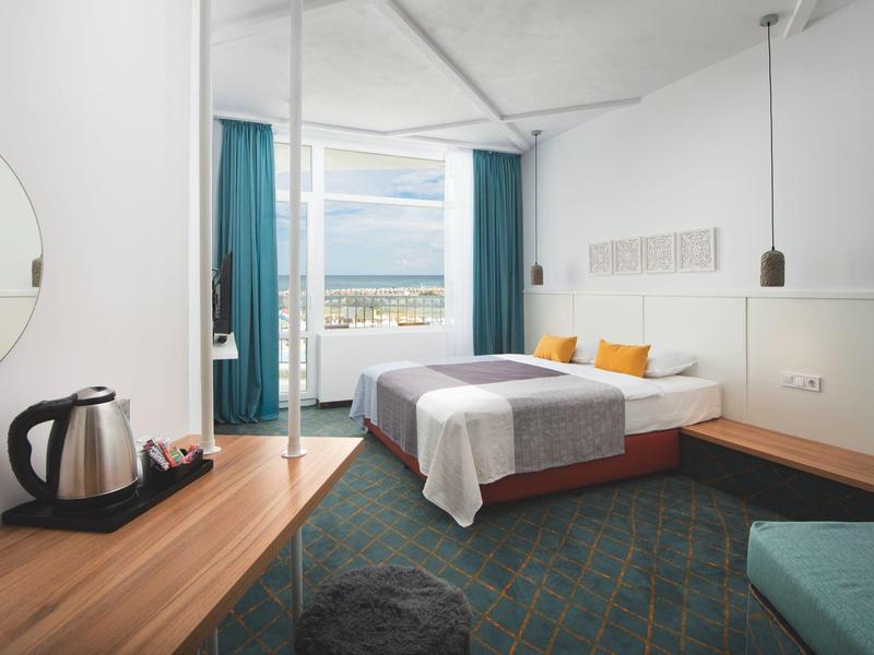 Moderner Hotelzimmer mit großem Fenster, Doppelbett, Holzmöbeln und blau-grünem Teppichboden.