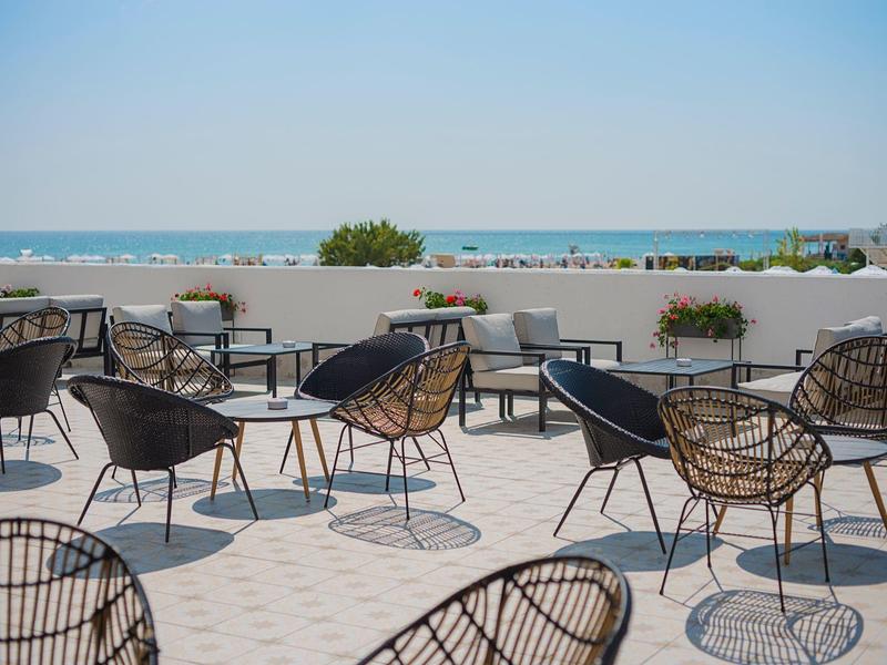 Leere Terrasse mit schwarzen Stühlen und Tischen, dahinter Meer und blauer Himmel.