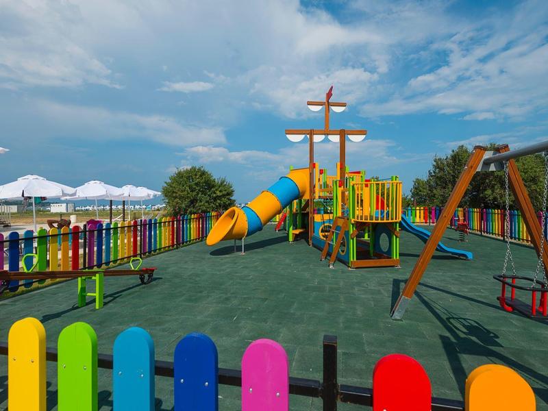 Bunter Spielplatz mit Rutsche, Schaukel und buntem Zaun unter blauem Himmel.