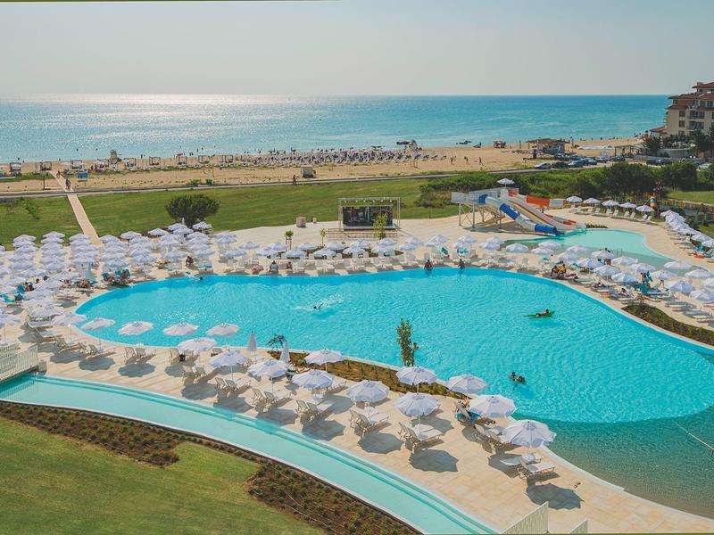 Großer Hotelpool mit weißen Sonnenschirmen und Blick auf das Meer im Hintergrund.