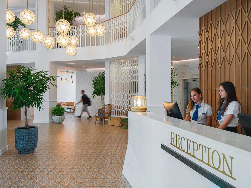 Moderne Hotellobby mit weißer Rezeption, warmen Holzelementen und Pflanzen in großen Töpfen.
